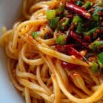 Close-up of spicy garlic noodles with chili oil, scallions, and red pepper flakes in a white bowl
