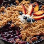 Blueberry Peach Crumble in a cast iron skillet with peach slices, blueberries, and whipped cream topping